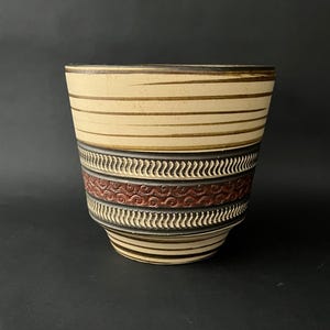 May include: A ceramic vase with a wide, cylindrical shape. The top section features horizontal beige stripes with brown accents. The middle band has a repeating pattern of red swirls, and the bottom section has a repeating pattern of white and black lines.