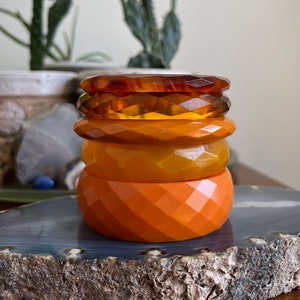 Puede incluir: Un conjunto de cuatro pulseras de plástico naranja y ámbar. Las pulseras son de diferentes tonos de naranja y tienen un patrón geométrico.