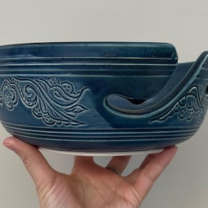 Puede incluir: Un cuenco de hilo de cerámica de color verde azulado con un patrón decorativo de flores y remolinos. El cuenco tiene un asa curva y una ranura para sujetar el hilo. El cuenco se sostiene en una mano.