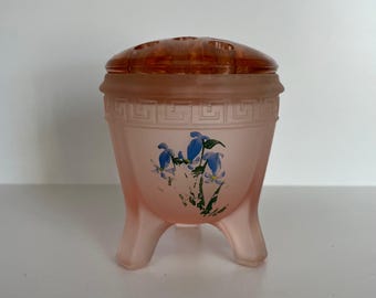 May include: A pink frosted glass vase with a floral design and a removable lid. The vase has three legs and a geometric pattern around the top.
