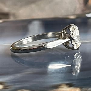 Puede incluir: Un anillo de compromiso de plata con un diamante redondo y transparente engastado en una montura de garras. El anillo est&aacute; sobre una superficie azul y gris.