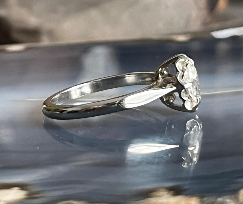 Puede incluir: Un anillo de compromiso de plata con un diamante redondo y transparente engastado en una montura de garras. El anillo est&aacute; sobre una superficie azul y gris.