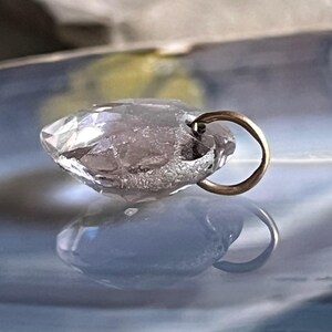 May include: A clear, faceted crystal pendant with a gold-colored bail. The pendant is teardrop-shaped and reflects light, creating a sparkling effect. The background is a blurred mix of blue and yellow.