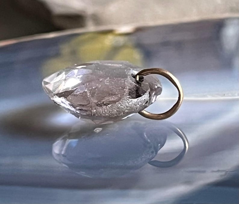 May include: A clear, faceted crystal pendant with a gold-colored bail. The pendant is teardrop-shaped and reflects light, creating a sparkling effect. The background is a blurred mix of blue and yellow.