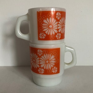 Puede incluir: Dos tazas de cerámica blanca con diseños florales naranjas. Las tazas están apiladas una encima de la otra.