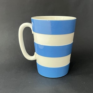 Puede incluir: Una taza de cerámica con un asa blanca y un cuerpo cilíndrico. La taza presenta bandas horizontales alternas de color azul claro y blanco. La taza está vacía y se encuentra sobre un fondo oscuro.