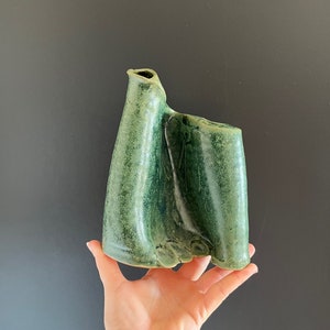 Puede incluir: Un jarrón de cerámica verde con una forma orgánica abstracta. El jarrón tiene una superficie texturizada y un diseño asimétrico único.