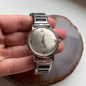 May include: A silver Benrus watch with a white face and black hands. The watch has a silver band and says "Selfwinding 17 Jewels" on the face.