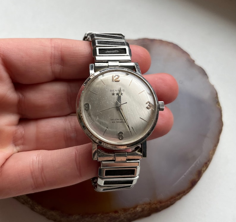 May include: A silver Benrus watch with a white face and black hands. The watch has a silver band and says "Selfwinding 17 Jewels" on the face.