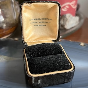 Pode incluir: Caixa de joias preta vintage aberta, com interior em cetim dourado e inserção em veludo preto. A caixa é estampada com um padrão geométrico e apresenta o texto "MITCHELL-COPP LTD. DIAMOND MERCHANTS WINNIPEG".