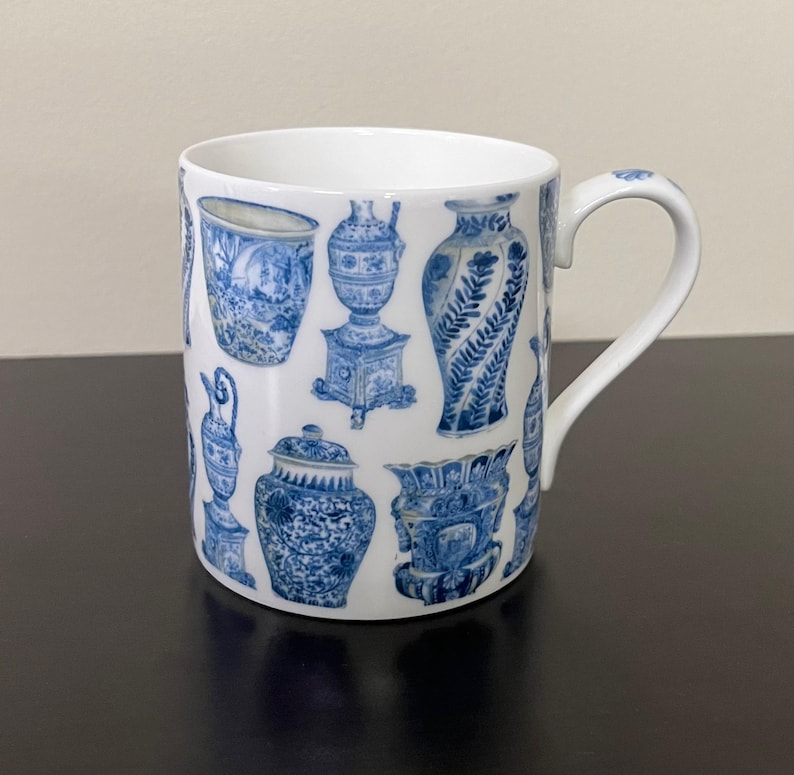 Puede incluir: Taza de cer&aacute;mica blanca con un patr&oacute;n repetido de jarrones antiguos azules y blancos. La taza tiene un asa curva y un dise&ntilde;o sencillo. Los patrones azules son detallados e intrincados, a&ntilde;adiendo un toque de estilo cl&aacute;sico.