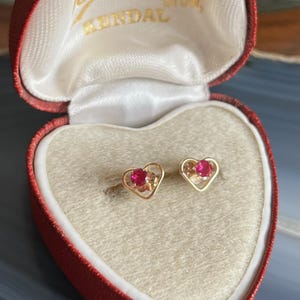 May include: Gold heart-shaped stud earrings, each with a small red gemstone, displayed in a red heart-shaped box. The box is open, revealing the earrings against a cream-coloured interior.