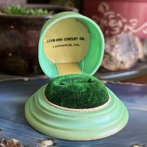 Puede incluir: Caja de joyería vintage verde claro abierta con un inserto de terciopelo verde. La caja es redonda con un diseño acanalado y una tapa con bisagras. El interior de la tapa tiene el texto "LEHM ANN JEWELRY CO. LaGrange, GA."