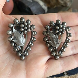 May include: A pair of silver tone clip-on earrings with a floral design. The earrings are shaped like a teardrop and feature a cluster of small beads around the edge.