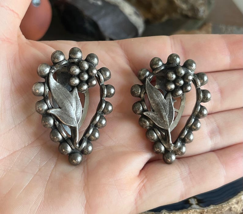 May include: A pair of silver tone clip-on earrings with a floral design. The earrings are shaped like a teardrop and feature a cluster of small beads around the edge.