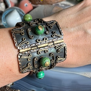 Op de afbeelding: Een brede, scharnierende manchetarmband met een donkere, getextureerde metalen afwerking. De armband heeft decoratieve messing accenten en groene, bolvormige stenen. Het ontwerp bevat gestileerde krul- en wervelpatronen.