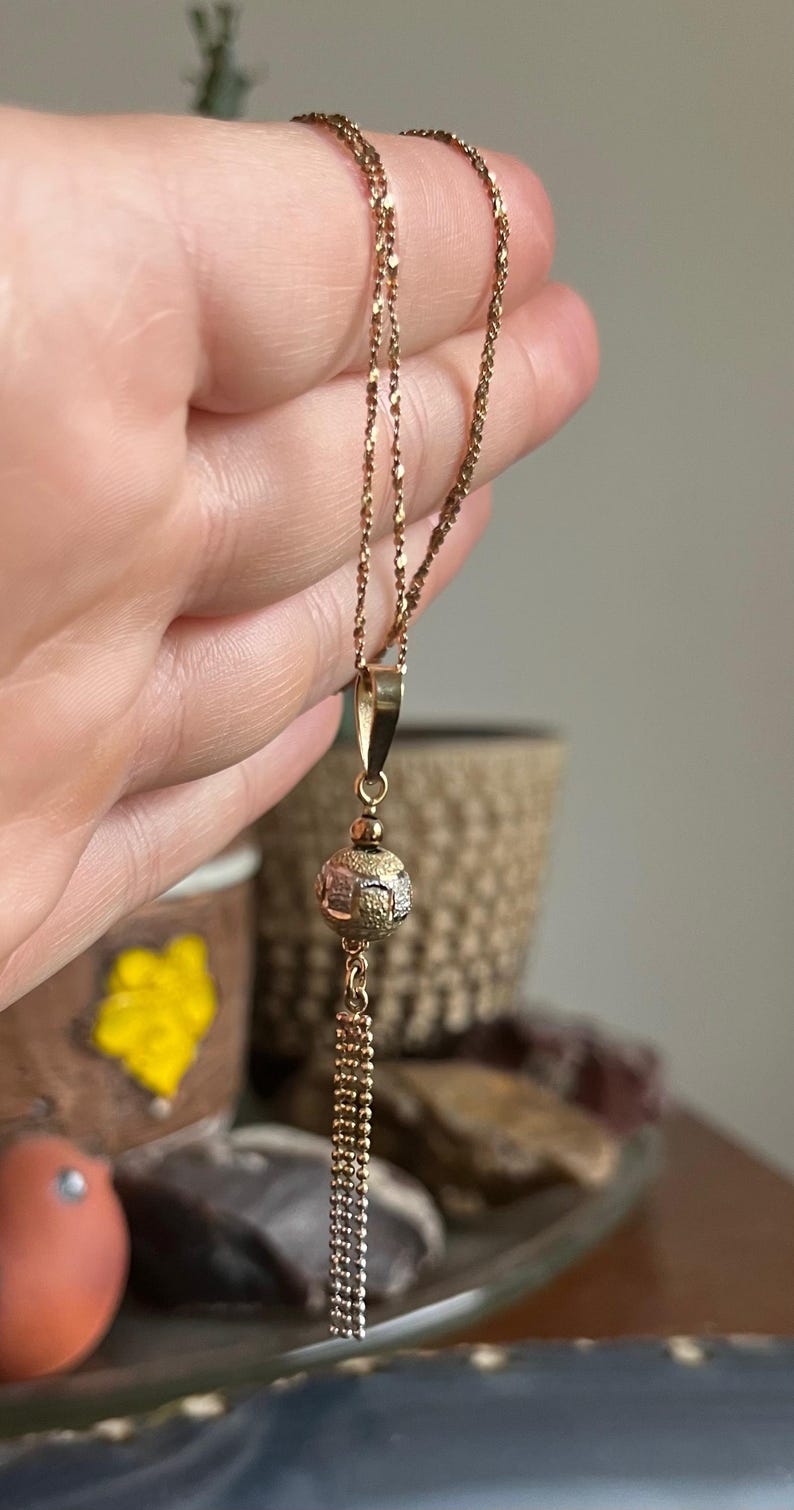Puede incluir: Un collar de cadena de oro con un colgante redondo con un dise&ntilde;o texturizado. El colgante est&aacute; unido a una cadena de borla.