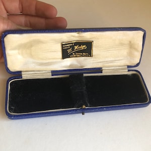 Op de afbeelding: Een blauwe fluwelen juwelendoos met een wit interieur en een gouden label met de tekst "Watchmaker & Jeweller W. Hodge 16 London South Campbelltown".