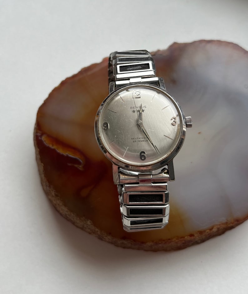 May include: A silver Benrus self-winding watch with a white face and black hands. The watch has a silver metal band with black accents.