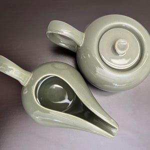 Könnte beinhalten: Eine salbeigrüne Keramik-Teekanne mit passendem Sahnekännchen. Die Teekanne hat einen runden Körper, einen Deckel mit kleinem Knopf und einen gebogenen Griff. Das Sahnekännchen hat eine längliche Form mit Ausguss und Griff. Auf dunkler Oberfläche.