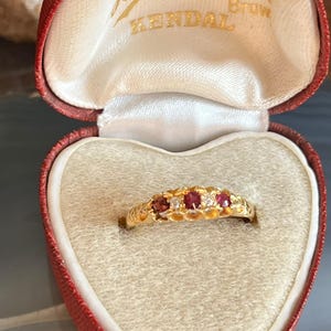 May include: A gold ring with a series of red and white gemstones, presented in a heart-shaped red jewelry box. The ring features a decorative, openwork design. The box has the text "H. Hoga" and "Kendal" printed on the inside.