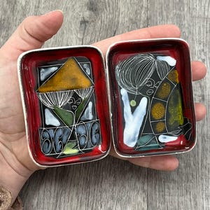 May include: Two rectangular, hinged enamel boxes with silver-colored metal edges. The boxes have a red border and black interior with abstract art in white, green, yellow, and brown. The boxes are open, revealing the artwork.