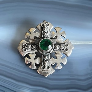 May include: A silver-toned cross pendant with an emerald green gemstone at the center. The cross features intricate floral and geometric detailing, with smaller crosses at each arm's end. The pendant is likely a brooch or a necklace charm.
