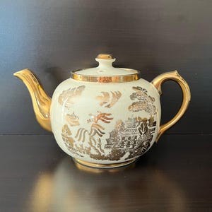May include: Cream-coloured teapot with gold accents. The teapot has a gold handle, spout, and lid trim. The body is decorated with a gold Asian-inspired landscape design. A classic piece for tea lovers.