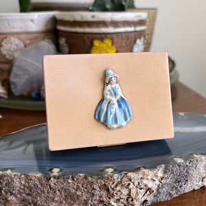 May include: A small, rectangular, pale pink box with a ceramic figurine of a girl wearing a blue dress and white hat. The box is sitting on a grey and brown agate slice.