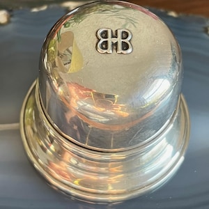 May include: A shiny silver bottle stopper with a domed top and a wide base. The top features a raised emblem with the intertwined letters "BB". The stopper reflects the surrounding environment, creating a mirrored effect.