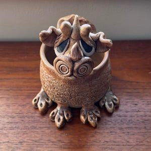 May include: A whimsical, tan-colored ceramic creature with large, expressive eyes and a textured body. The open-mouthed figure has a bowl-like interior, and is perched on four clawed feet. The creature has a horn and ear-like protrusions.
