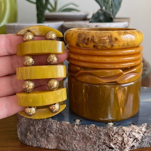 May include: A set of four yellow and brown plastic bangles. The bangles are stacked on top of each other. The top bangle has a marbled pattern. The other bangles are solid colours. The bangles are decorated with gold-coloured beads.