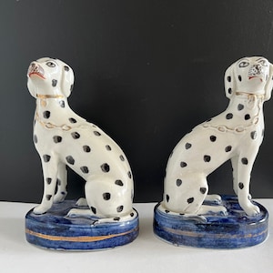 Op de afbeelding: Twee keramische dalmatiërhondjes met zwarte vlekken op een witte achtergrond. De honden zitten op blauwe en gouden sokkels.