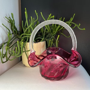 Pode incluir: Uma pequena cesta de vidro rosa com uma alça de arame prateado. A cesta está sobre uma superfície branca ao lado de uma planta em vaso.