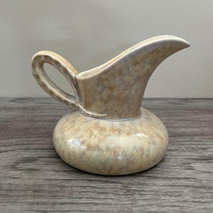 Könnte beinhalten: Ein kleiner Keramikkrug mit rundem Boden und gebogenem Ausguss. Der Krug hat einen Henkel und ist mit einer gesprenkelten, marmorierten Glasur aus Beige, Hellbraun und Hellbraun versehen. Der Krug ist wahrscheinlich Vintage oder antik.