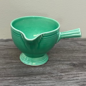 May include: A turquoise gravy boat with a handle and a spout. The boat has a round bowl shape and sits on a pedestal base. The handle is ribbed, and the bowl has horizontal ridges. The gravy boat is made of ceramic.