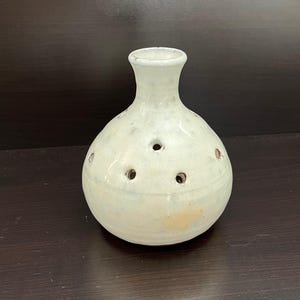 May include: A small, off-white ceramic vase with a rounded body and a narrow neck. The vase has several small, evenly spaced holes around its body. The surface has a slightly textured appearance.