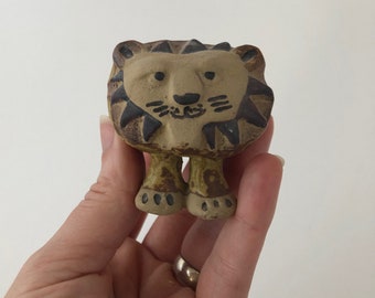 May include: A small ceramic lion figurine with brown and black paint. The lion has a simple, cartoon-like face with black eyes and a black nose.