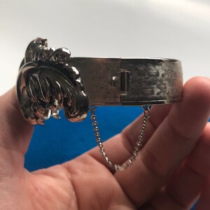 K&ouml;nnte beinhalten: Silberner Scharnier-Armreif mit floralem Design und einer Kette an jeder Seite.