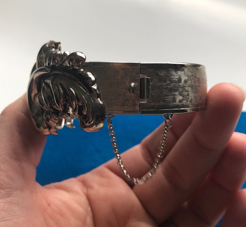 K&ouml;nnte beinhalten: Silberner Scharnier-Armreif mit floralem Design und einer Kette an jeder Seite.