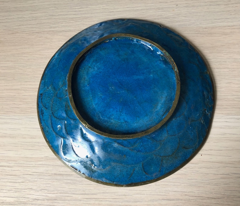 May include: A blue ceramic plate with a brass rim and a decorative design. The plate has a raised rim and a smooth surface.