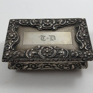 May include: A silver box with an ornate floral design and a monogrammed inscription that reads "T + D".