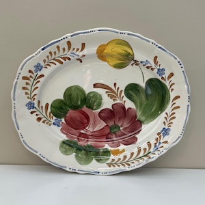 Könnte beinhalten: Ovale, weiße Keramikplatte mit gewelltem Rand und handgemaltem Blumendesign. Die Blumen sind in Rot-, Gelb- und Grüntönen gehalten, mit braunen und blauen Akzenten. Der Rand hat einen blauen Rand.