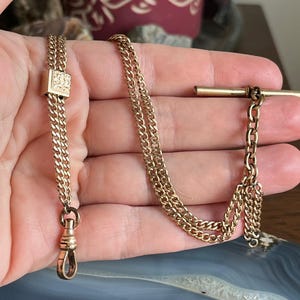 May include: A gold-coloured pocket watch chain featuring a T-bar and a clasp. The chain is made up of multiple linked sections, with a decorative square element near the clasp. The chain is held against a blue and grey surface.