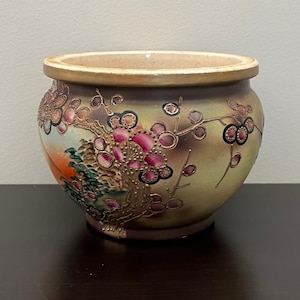 May include: A ceramic planter with a wide opening and a rounded body. The planter features a hand-painted design of a flowering tree with pink blossoms and gold accents. The background has a sunset scene with green foliage.