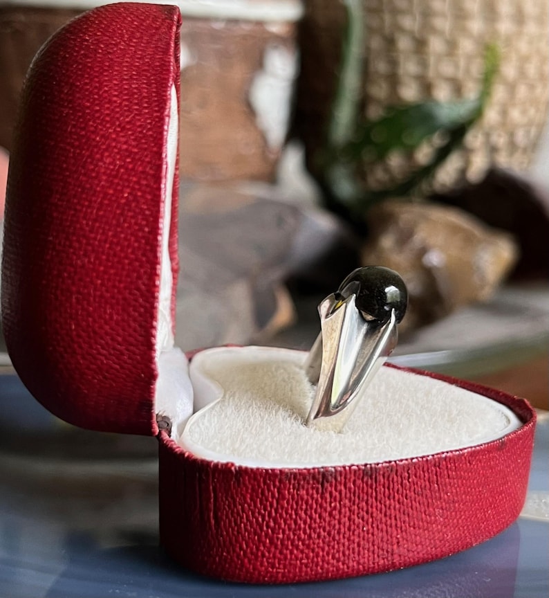 May include: A silver ring with a dark, round gemstone, presented in an open red heart-shaped box with a white interior. The ring's band has a unique, angular design.