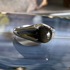 May include: A silver ring with a dark, speckled gemstone. The ring has a modern design with a polished finish. The gemstone is round and appears to have a reflective, golden sheen. The ring is set on a reflective surface.