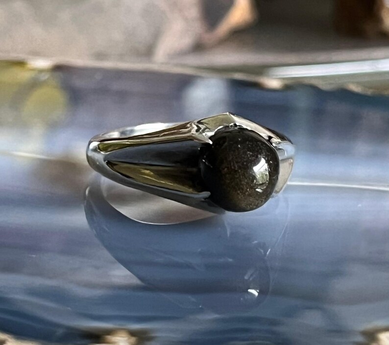 May include: A silver ring with a dark, speckled gemstone. The ring has a modern design with a polished finish. The gemstone is round and appears to have a reflective, golden sheen. The ring is set on a reflective surface.