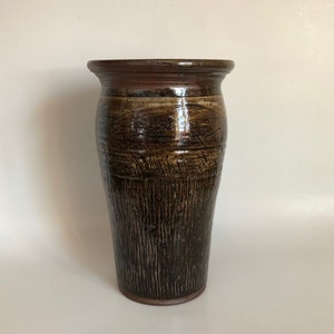 May include: A brown ceramic vase with a textured surface and a slightly flared opening. The vase has a dark brown glaze with streaks of lighter brown and a hint of red.