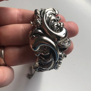 K&ouml;nnte beinhalten: Ein silbernes Armband mit einem verzierten, wirbelnden Design. Das Armband ist offen und hat einen Kettenverschluss.